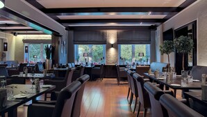 A modern dining area with multiple tables and chairs, large windows with views of greenery, and a central light fixture. - Landhaus Begatal (Doerentrup)