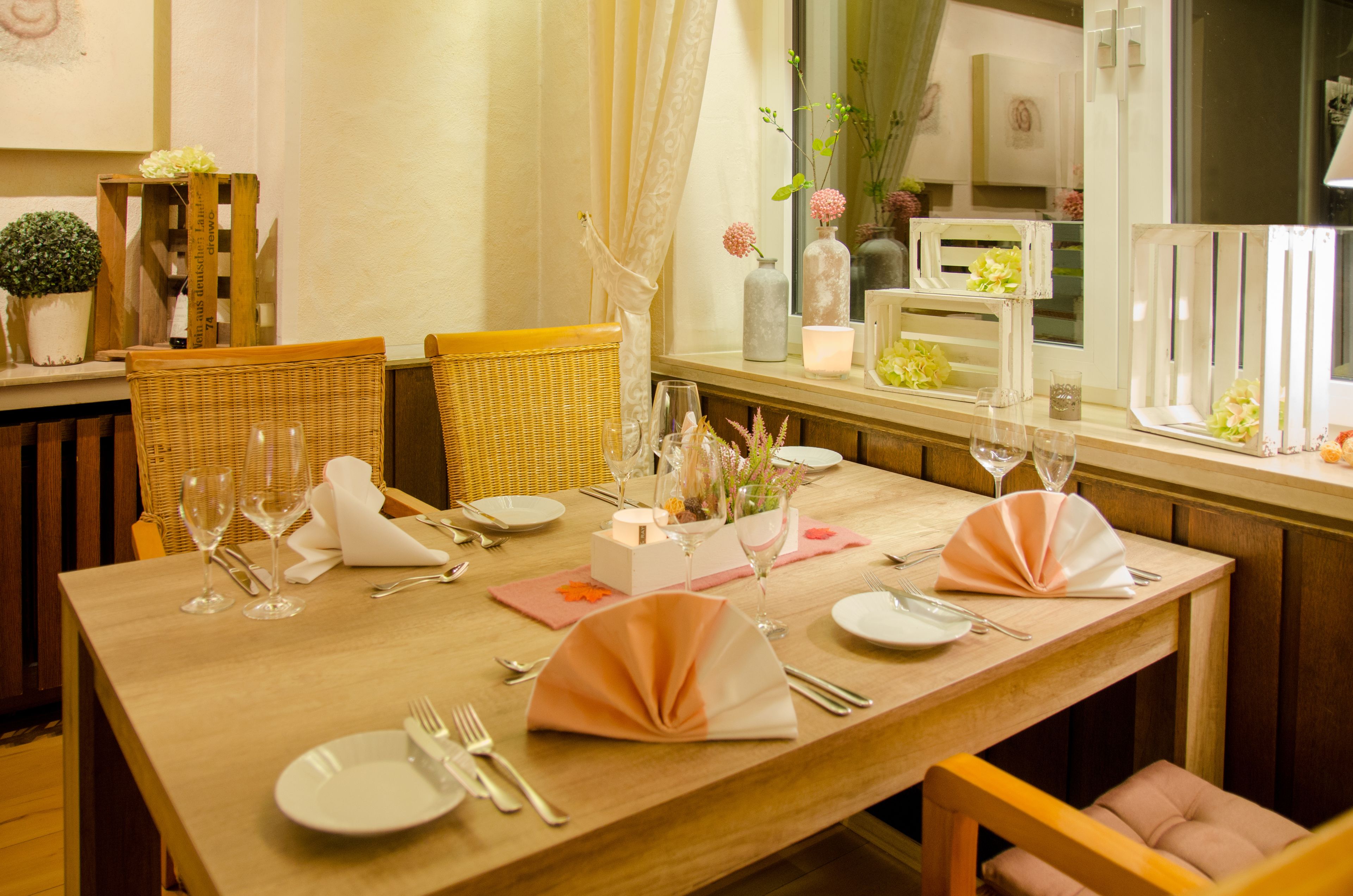 Una mesa de comedor dispuesta con platos blancos, cubiertos y servilletas rosas, rodeada de sillas de mimbre.