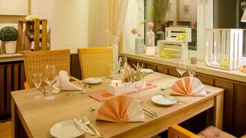 Una mesa de comedor dispuesta con platos blancos, cubiertos y servilletas rosas, rodeada de sillas de mimbre.