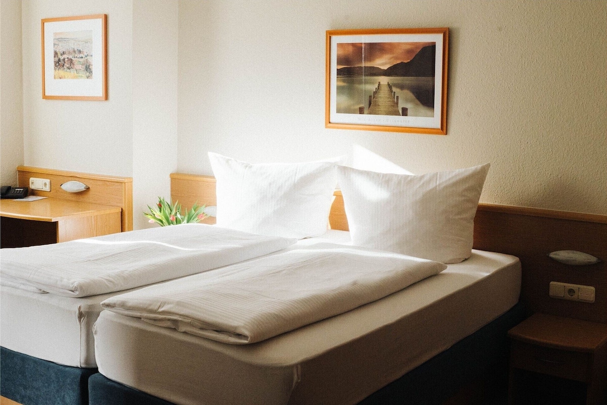A neatly made bed with white linens, a wooden headboard, and a bedside table with a lamp. There are two framed pictures on the wall.