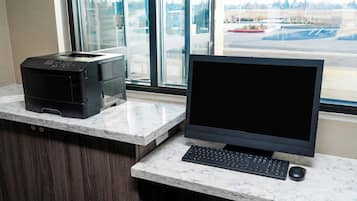 Un ordinateur de bureau installé sur un comptoir en marbre, avec une imprimante et un clavier à proximité, près d’une fenêtre donnant sur un parking.
