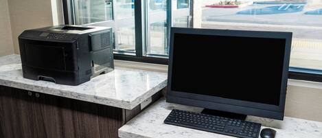 A desktop computer setup with a printer and keyboard on a marble countertop, near a window with a view of a parking lot.
