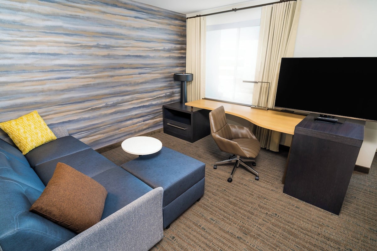 Une chambre d’hôtel moderne comprenant un canapé d’angle, un bureau avec un téléviseur à écran plat, une petite table ronde et une grande fenêtre avec des rideaux.