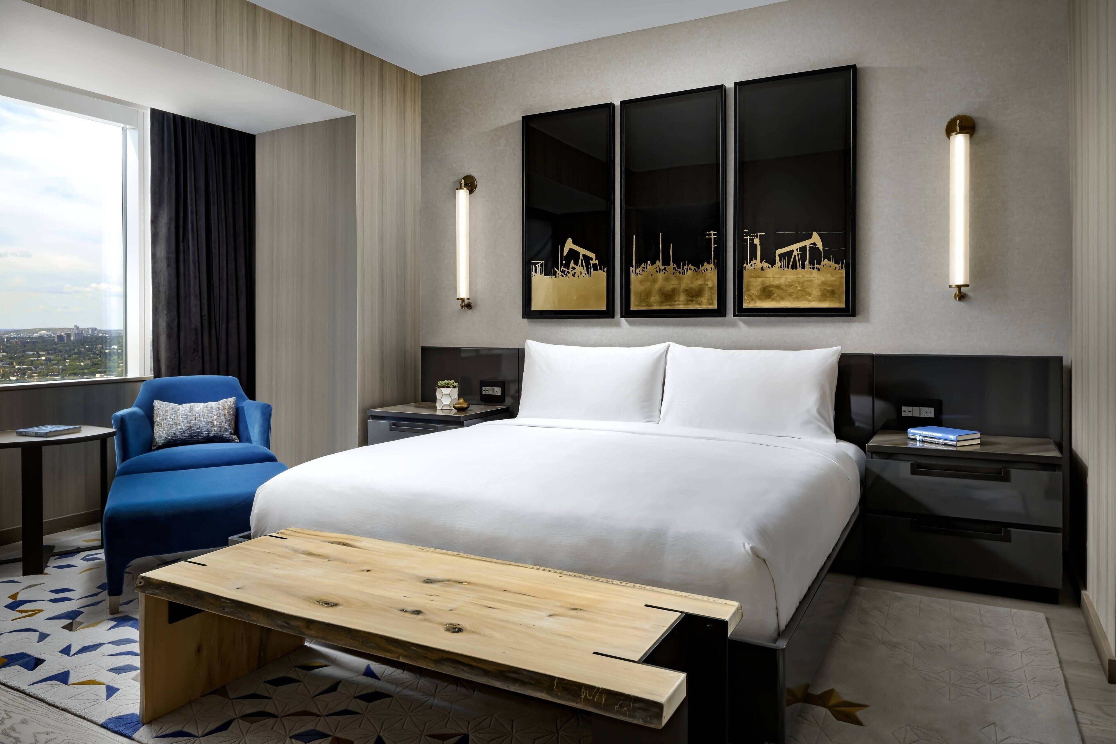 Una camera d'hotel moderna con un letto grande, una panca in legno, una poltrona blu e vista sulla città.