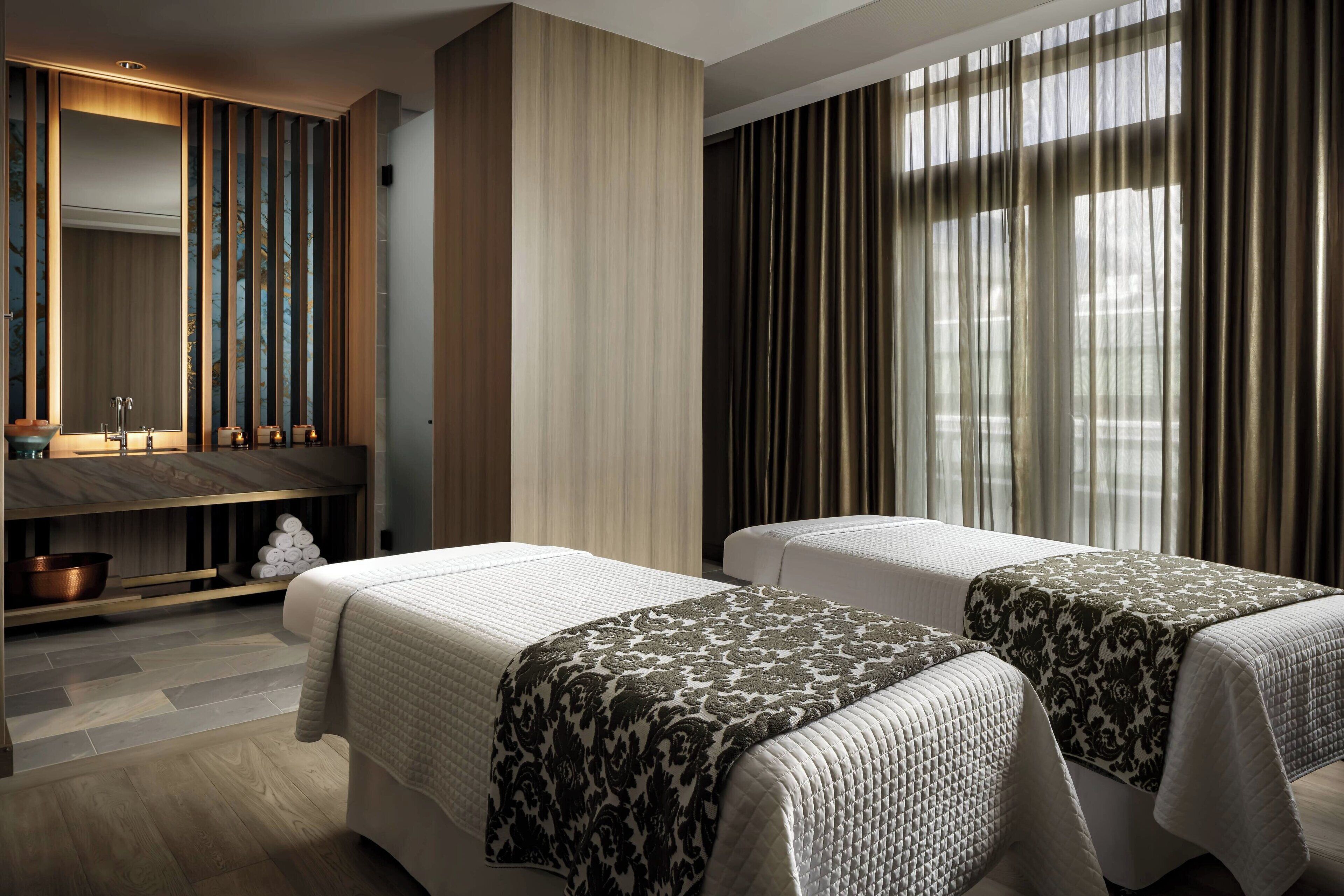 Habitación de hotel con dos camas, un tocador con espejo, un lavabo y un amplio ventanal con cortinas.