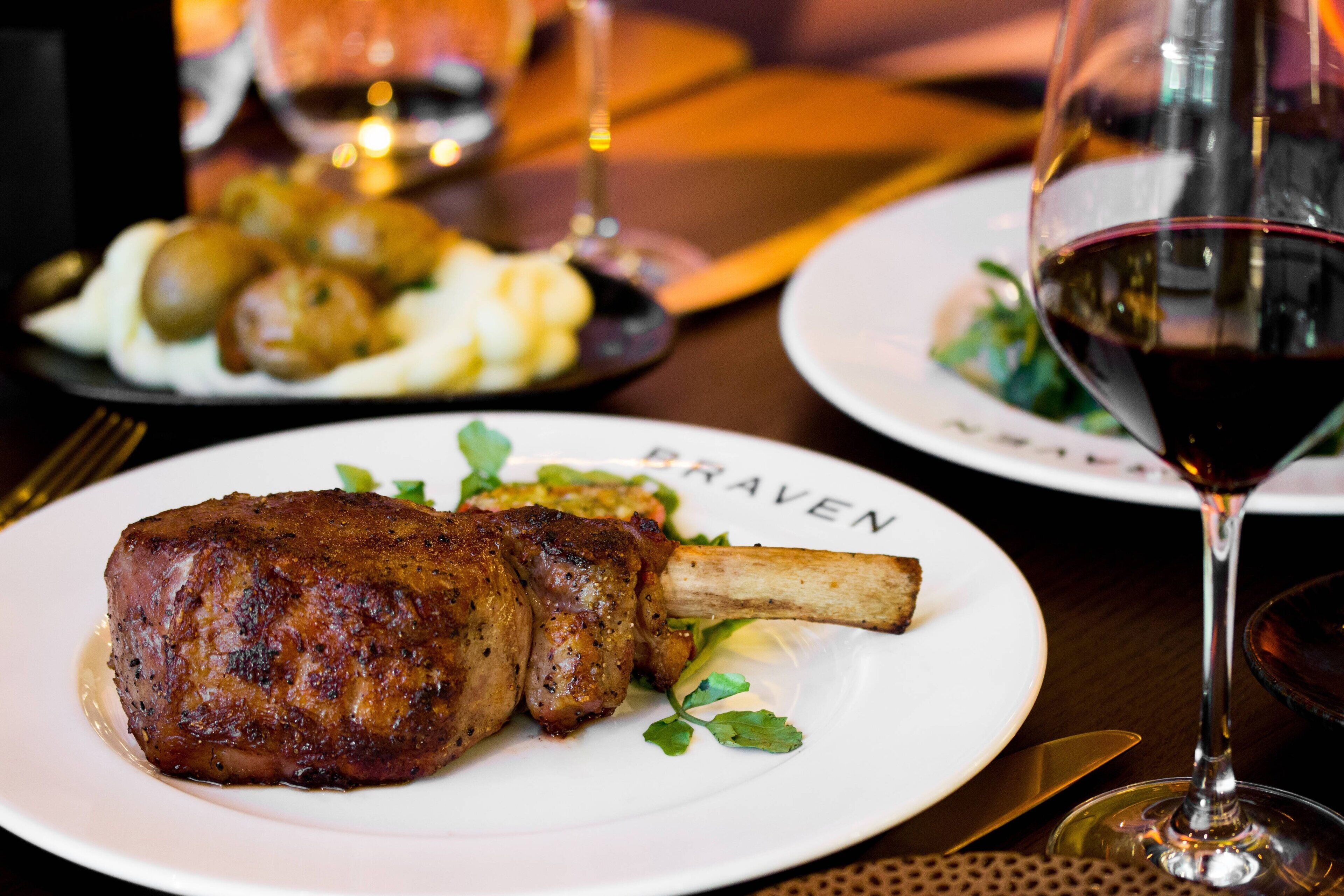 Une assiette de nourriture comprenant un jarret d’agneau et des légumes verts, un verre de vin rouge, et une autre assiette avec des pommes de terre et des légumes verts en arrière-plan.