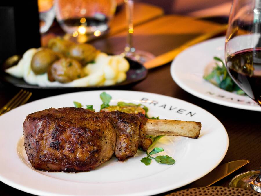 Prato com um ensopado de cordeiro e vegetais, uma taça de vinho tinto, e outro prato com batatas e vegetais ao fundo.