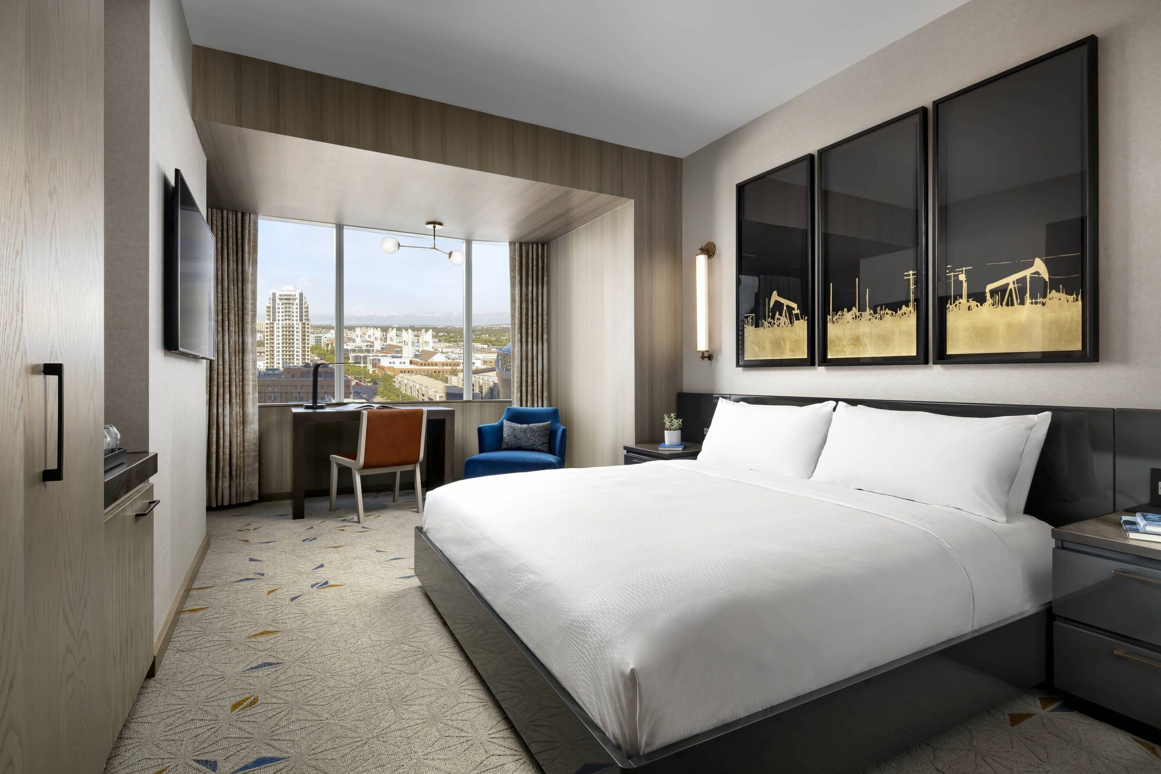 A modern hotel room with a large bed, a desk, and a chair. There is a large window offering a city view.