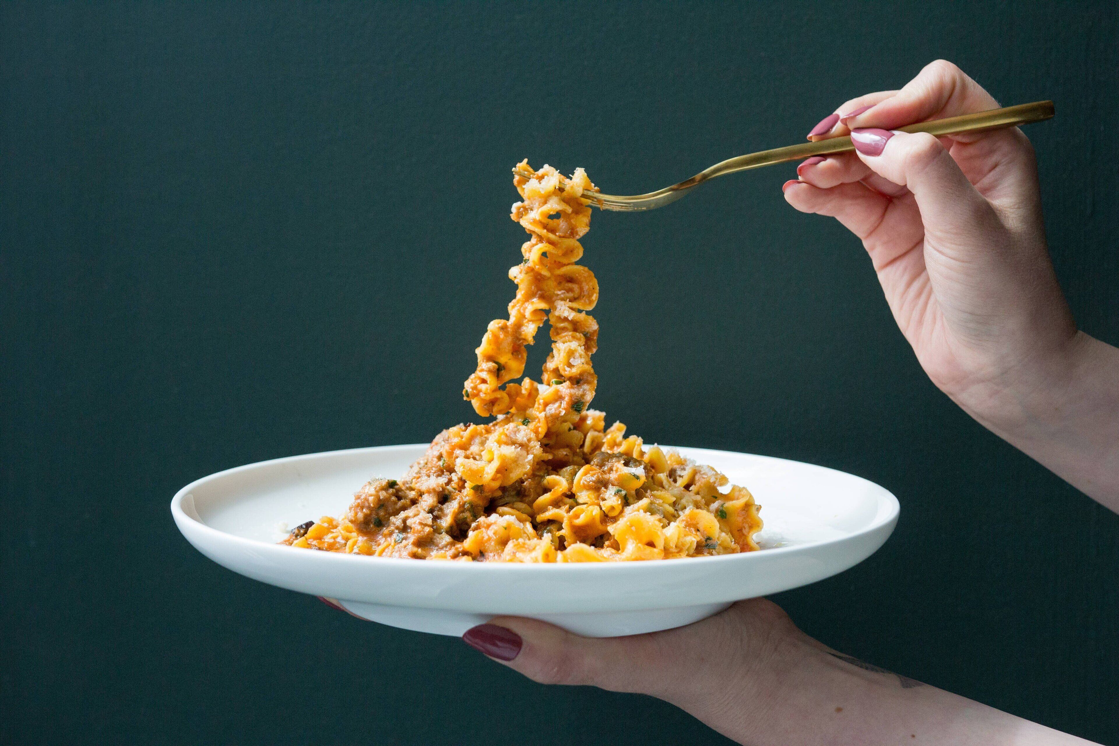 A hand holding a plate with pasta and a fork lifting some pasta.