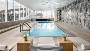 An indoor swimming pool with clear glass walls, a marble wall with a geometric pattern, and a wooden bench with a towel.