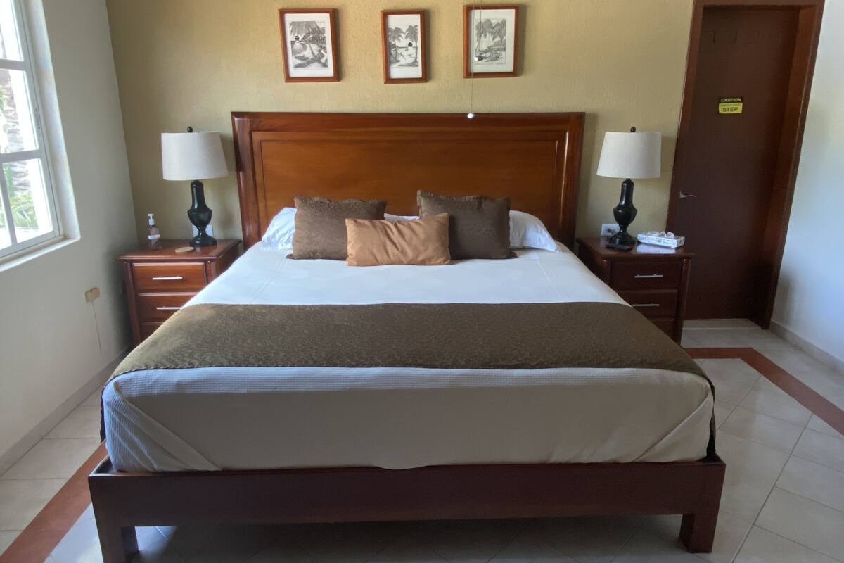 Una cama bien hecha con cabecera de madera, dos mesitas de noche con lámparas, y tres cuadros enmarcados en la pared.