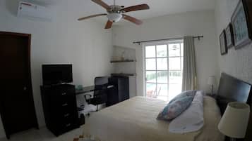 Ein Schlafzimmer mit Bett, Schreibtisch, Stuhl, Fernseher, Deckenventilator und einem Fenster mit Vorhängen.