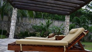 A wooden lounge chair with a cushion under a pergola, set against a stone wall and lush greenery. - YurInn (Cancun)