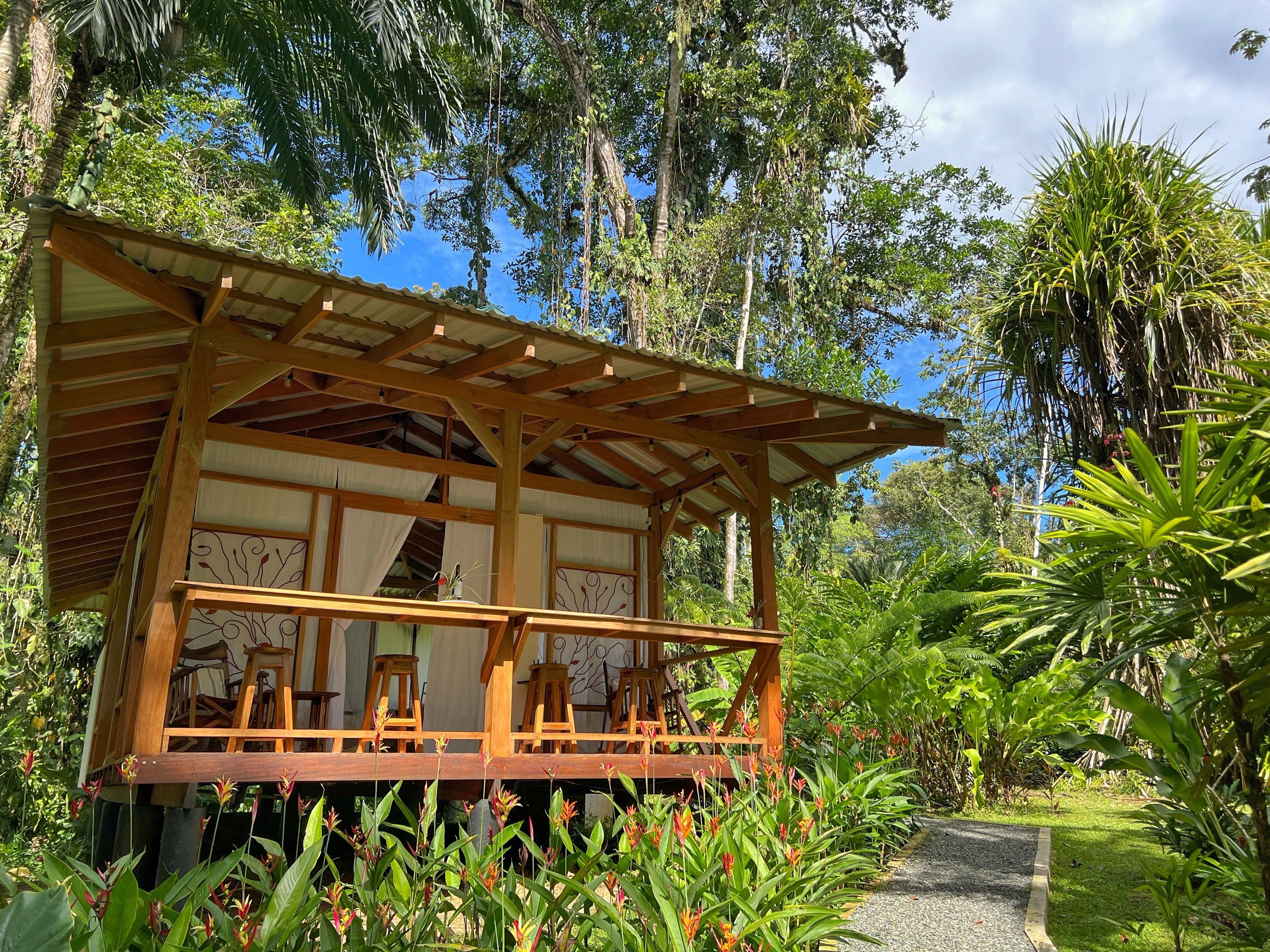 Una struttura in legno con un portico coperto, circondata da vegetazione lussureggiante e piante tropicali.