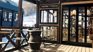 A wooden deck with outdoor seating, a large window, and a view of a snowy landscape. - ALP Lodge (Hakuba)