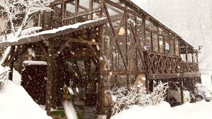 A wooden building with a sloped roof, surrounded by snow-covered trees and bushes. - ALP Lodge (Hakuba)
