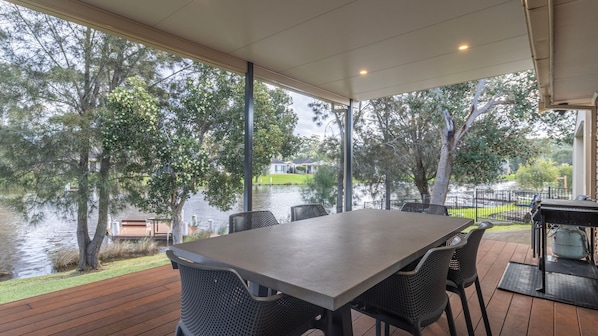 Un espace extérieur abrité, aménagé pour prendre les repas, avec une table et des chaises, donnant sur un lac bordé d’arbres et sur une maison située au loin.