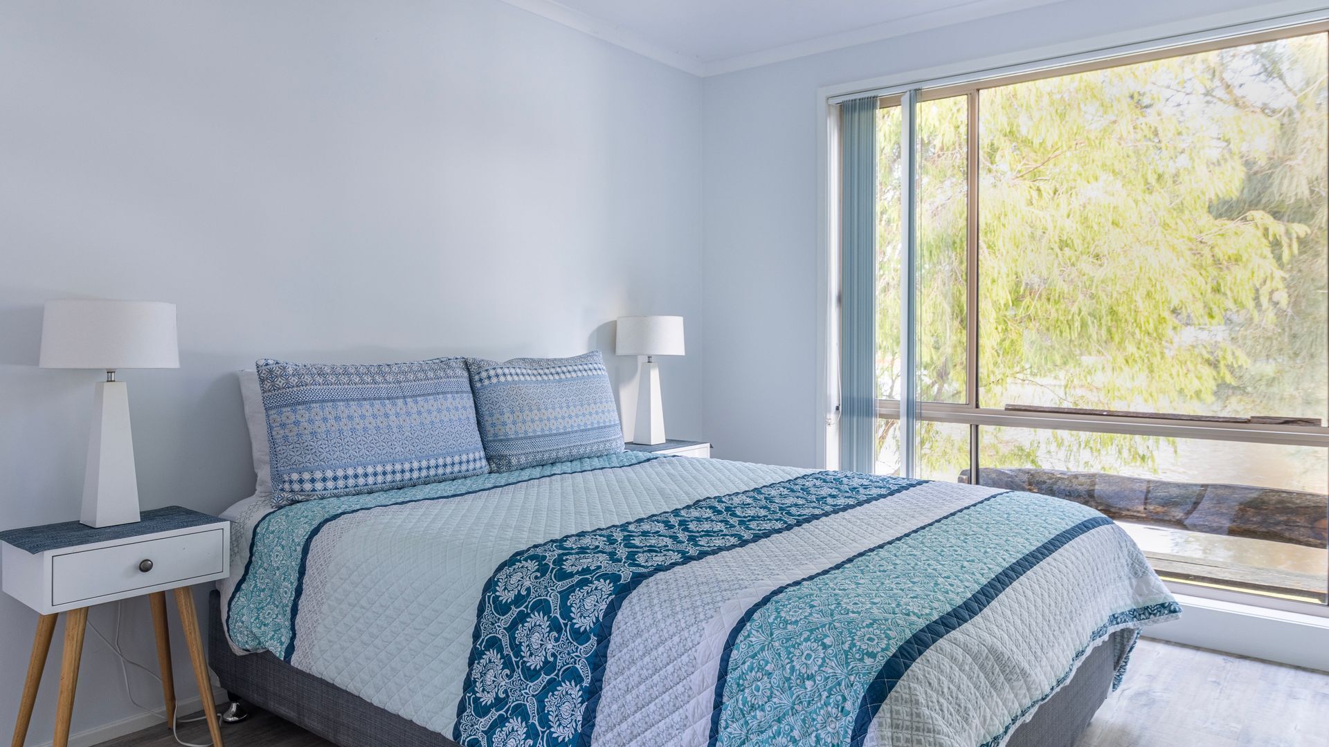 A bedroom with a large window, a bed with patterned bedding, two bedside tables with lamps, and a nightstand with a drawer.
