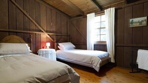 A room with two beds, wooden walls, a window with curtains, and a small table with a lamp.