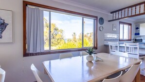 A dining area with a large window, white chairs, a wooden table, and a view of trees and sky. - Berrara Cove Beach House (Berrara)