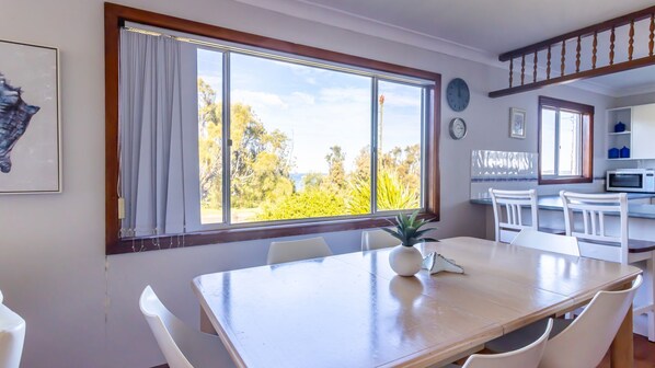 A dining area with a large window, white chairs, a wooden table, and a view of trees and sky. - Berrara Cove Beach House (Berrara)