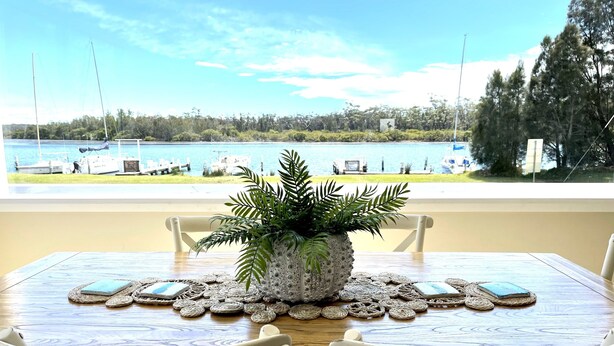 A dining table with a potted plant centerpiece, surrounded by white chairs, overlooking a lake with boats and greenery. - Riverbank Cottage - Waterfront (Sussex Inlet)