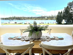 Una mesa de comedor con un centro de mesa de planta en maceta, rodeada de sillas blancas, con vista a un lago con botes y vegetación.