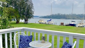 A wooden deck with two white metal chairs and a small round table, overlooking a calm lake with boats. - Riverbank Cottage - Waterfront (Sussex Inlet)