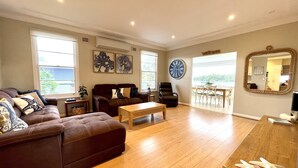 A spacious living room with a brown leather sofa, a wooden coffee table, and a dining area with a large clock and a mirror. - Riverbank Cottage - Waterfront (Sussex Inlet)