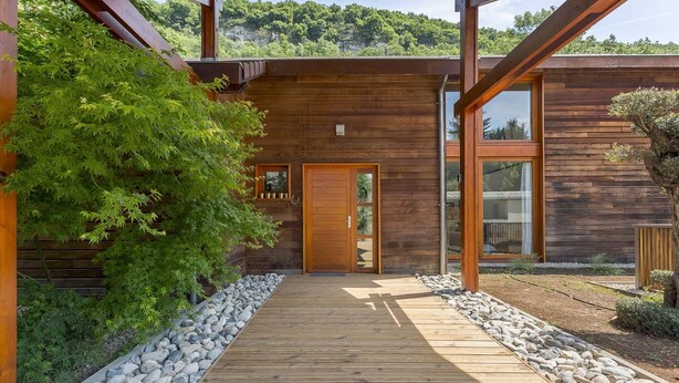 Una construcción de madera con un balcón de madera que lleva a una puerta, rodeada de follaje y rocas.
