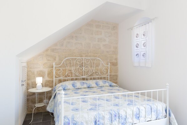 A bedroom with a sloped ceiling, a stone wall, a white metal bed frame, a bedside table with a lamp, and a window with a curtain. - Emotions of Casa Barone, villa with swimming pool, sea view, suitable for large groups (Scicli)