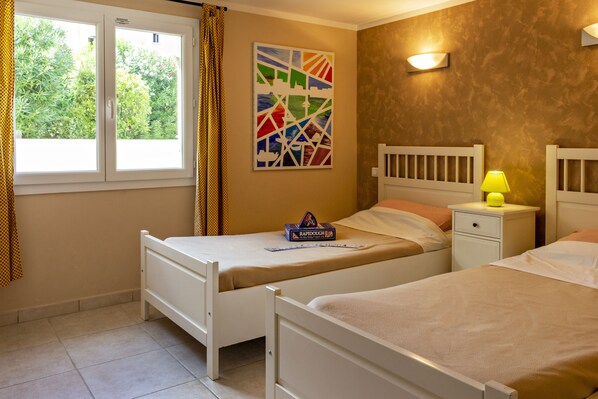 A bedroom with two single beds, a window with curtains, a wall-mounted artwork, a bedside table with a lamp, and a box of RapidDough on one bed.