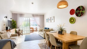 Un salon moderne comprenant un canapé, une table basse, une table à manger et des chaises. On y trouve également un téléviseur, une plante et une fenêtre avec des rideaux.