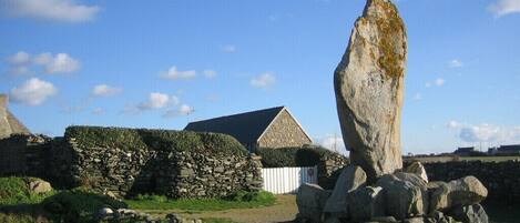 A stone structure with a large upright stone in the foreground, a stone wall, and a building with a sloped roof in the background.