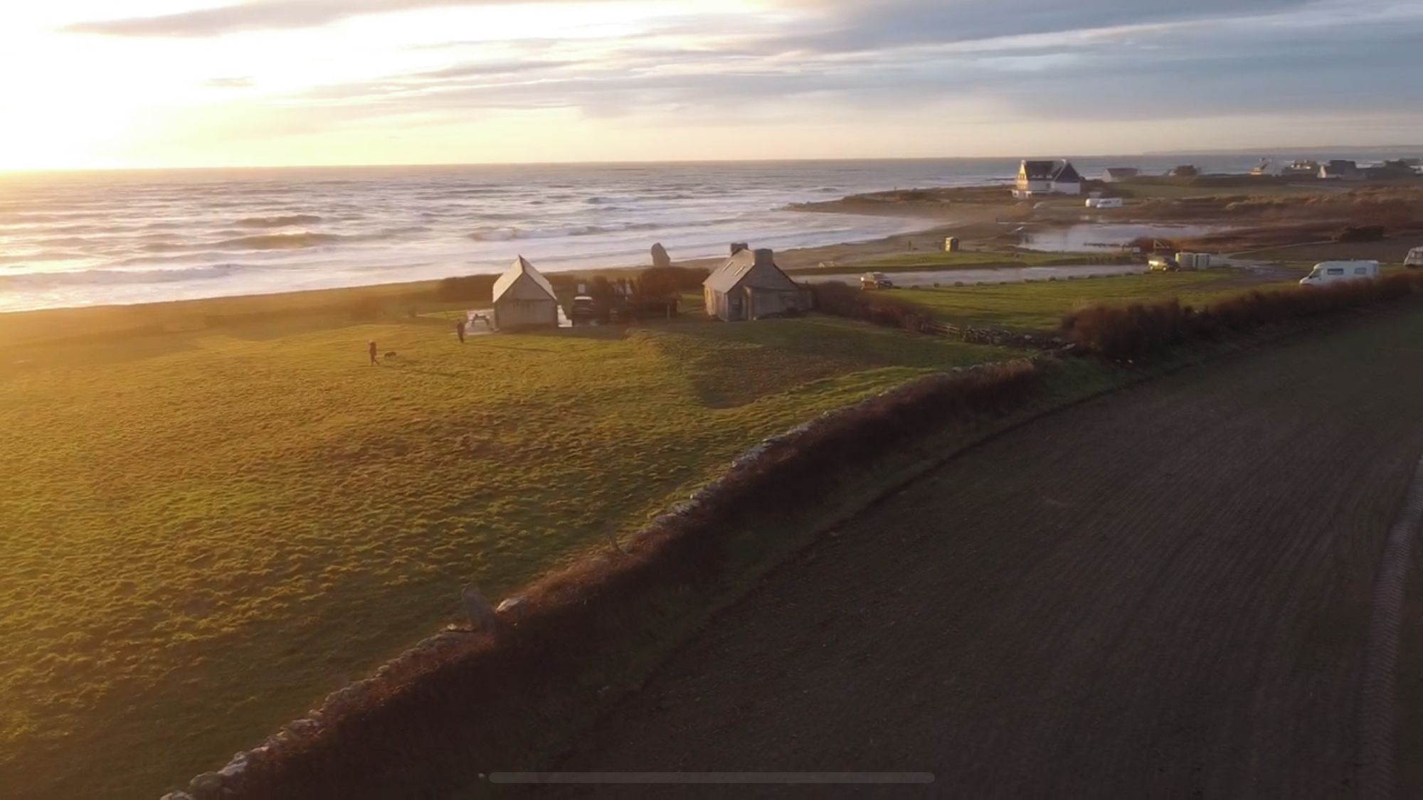 A coastal landscape with a grassy area, a few small buildings, and a body of water.