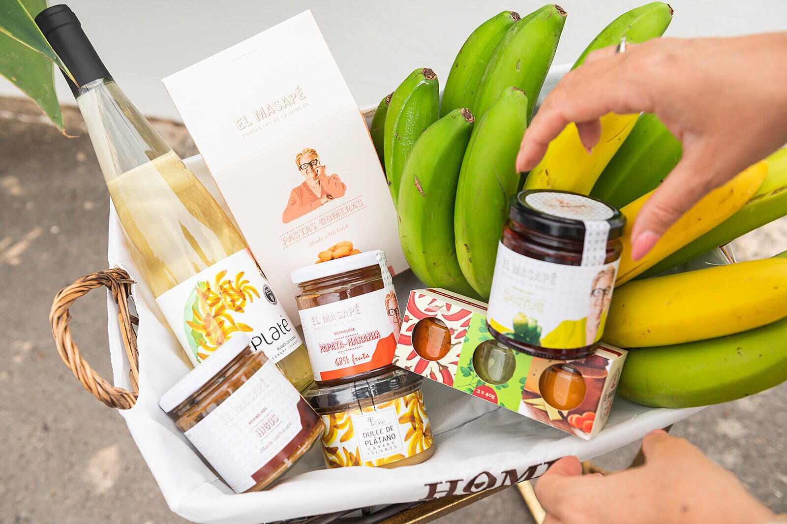 Un panier contenant une bouteille de vin blanc, une carte illustrée avec la photo d’un chef, des pots de confiture et une grappe de bananes.