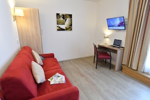 Ein Zimmer mit einem roten Sofa, einem Schreibtisch mit einem Laptop, einem Fernseher und einem Bild an der Wand.