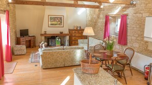 Un salon chaleureux avec une cheminée, des murs en pierre, des poutres apparentes, un coin repas avec une table et des chaises, et un ensemble de canapés.