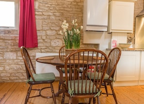 A wooden dining table with four chairs, a basket of flowers, and a white cabinet in a kitchen with stone walls.