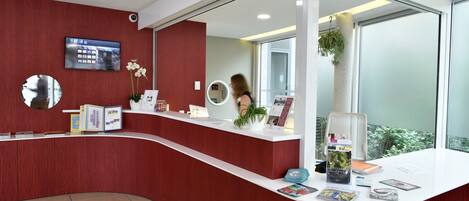 A reception desk with a red wall, a mounted TV, a mirror, and a window with a view of greenery.