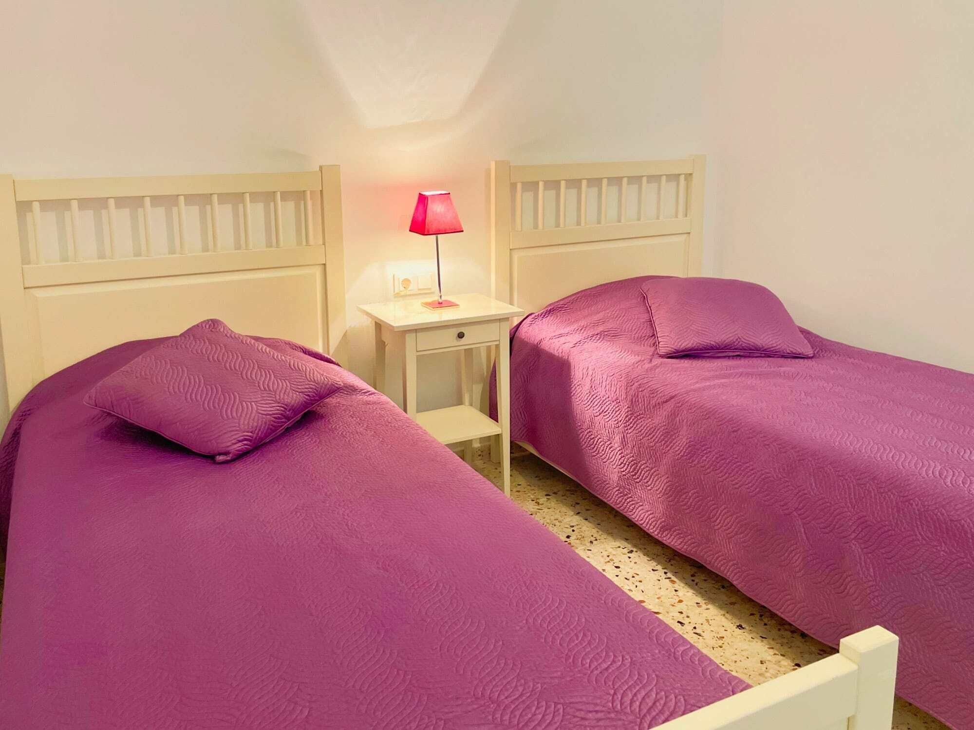 Una stanza con due letti, ciascuno con una coperta viola e cuscini abbinati. Tra i letti, un comodino con una lampada.