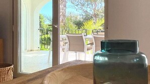 Une table en bois avec un bocal en verre au centre, un panier en osier à gauche, et, à travers une porte vitrée, on aperçoit un patio extérieur avec des chaises et des plantes.