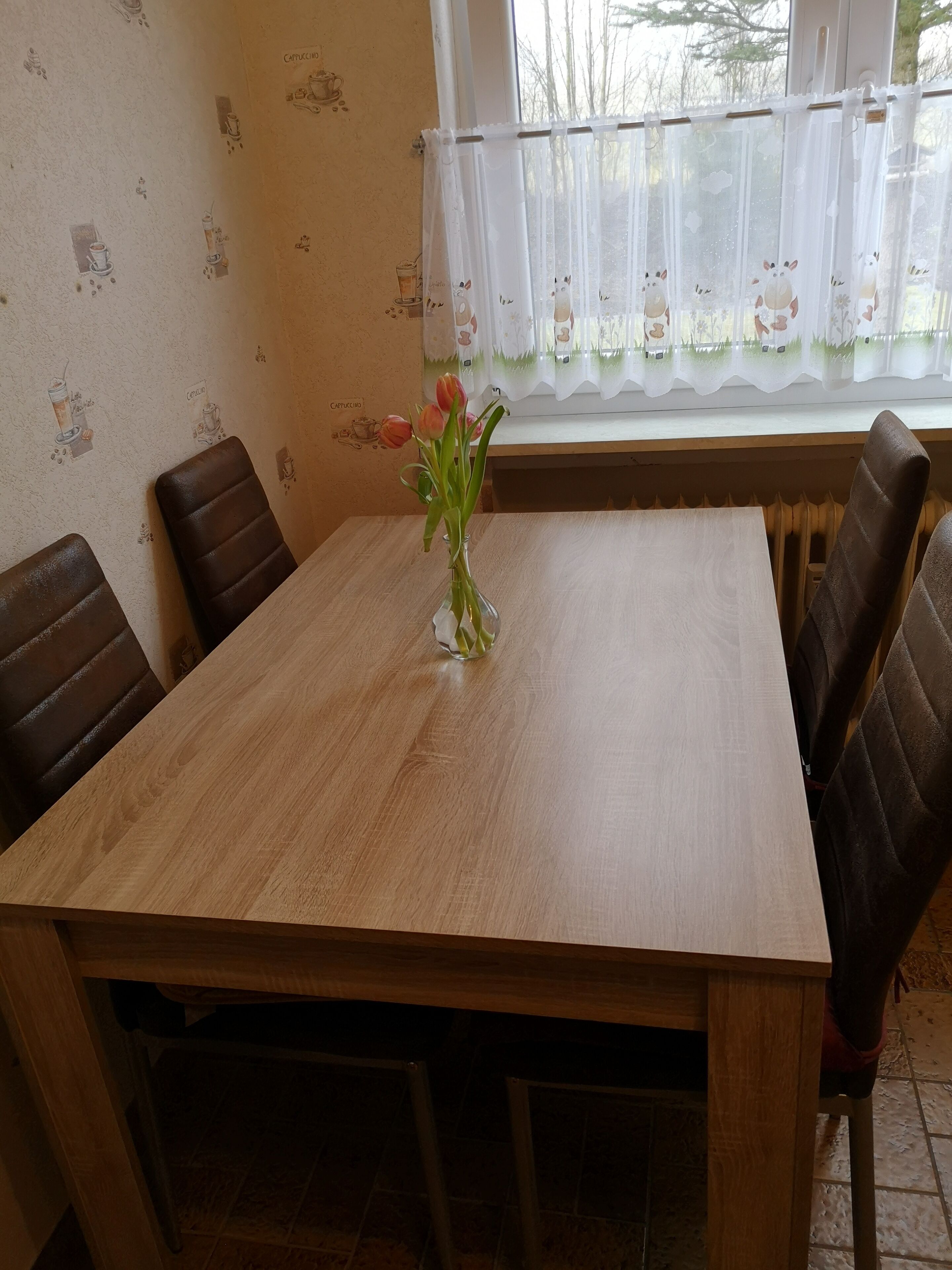 Une table à manger en bois, quatre chaises, un vase avec des fleurs et une fenêtre avec un rideau.