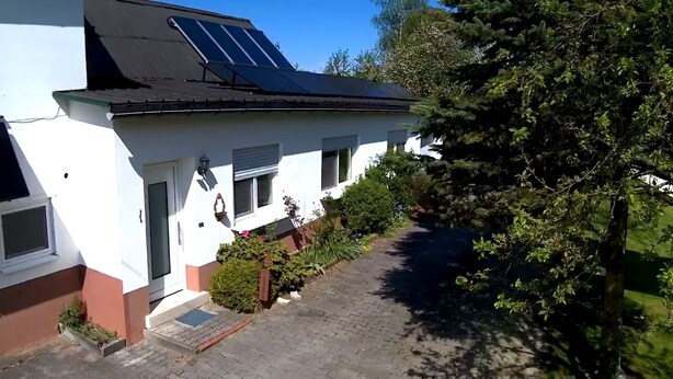 Uma casa branca com painéis solares no telhado, uma entrada pavimentada e um jardim com diversas plantas.