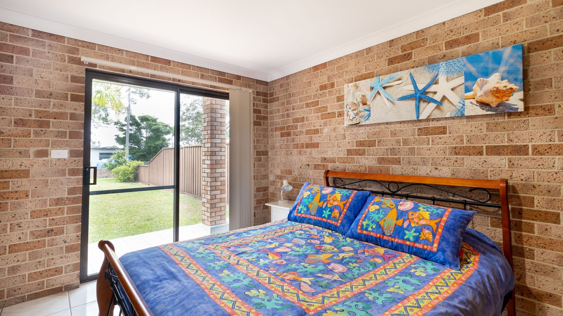 Une chambre avec un mur en briques, un lit recouvert d’une couette aux couleurs vives et une grande fenêtre donnant sur l’extérieur.