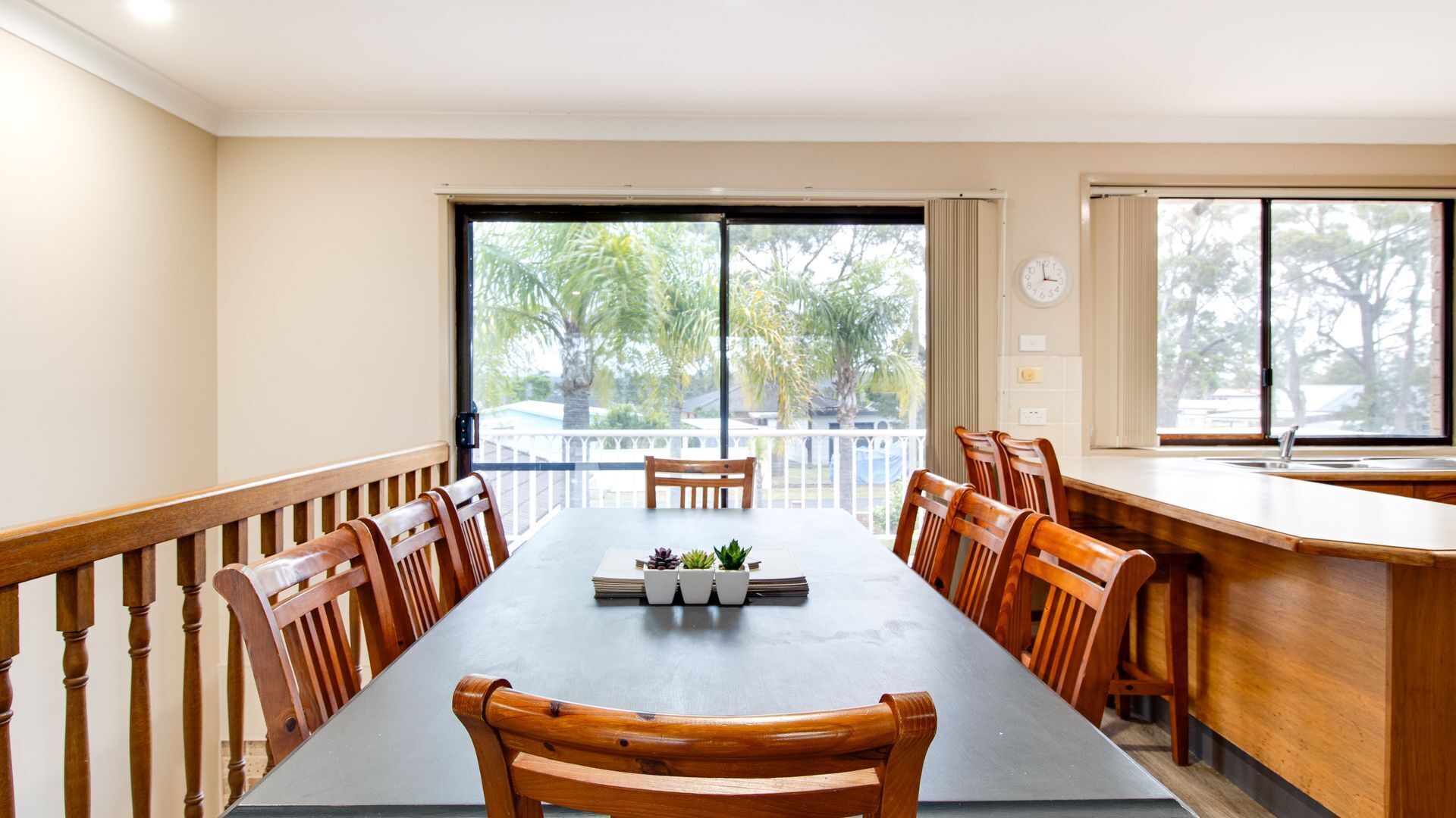 A dining area with a long table, wooden chairs, a small potted plant, and a view of trees through large windows.
