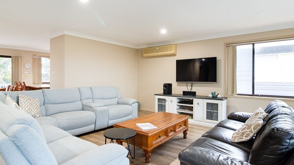 A living room with a light blue sofa, a black leather sofa, a wooden coffee table, and a TV on a white cabinet. - Hill Top Water and Bush Views (Sussex Inlet)