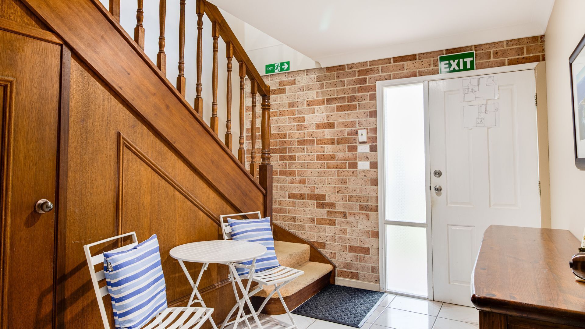 A hotel lobby with a wooden staircase, a white door with an exit sign, a small table with chairs, and a brick wall.