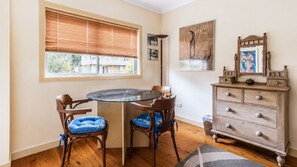 Una habitación con una mesa redonda de vidrio, dos sillas de madera con cojines azules, un aparador de madera con cajones, un espejo y obras de arte en la pared.