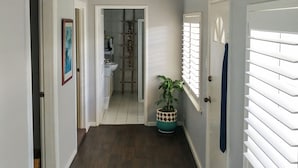 A hallway with wooden flooring, white walls, and a ceiling light. There are doors on both sides, a plant in a decorative pot, and a window with blinds. - Airport B&B (Redcliffe)